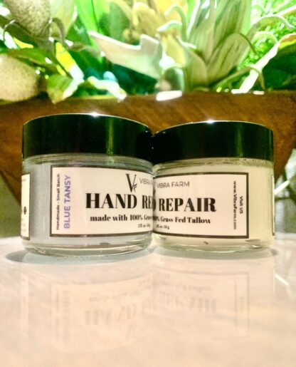 Vibra Farm Hand Repair Cream with Blue Tansy and a container of plain hand repair cream on a white marble surface with house plants in the background.