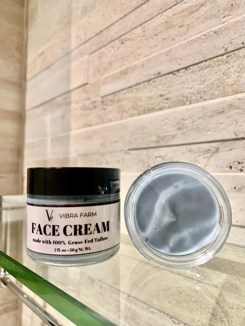 Image shows the Blue Tansy Oil, Honey, Jojoba and Vitamin E Whipped Tallow Face Cream from Vibra Farm on a glass shelf in a beautiful bathroom with an open container showing the blue color of the cream.